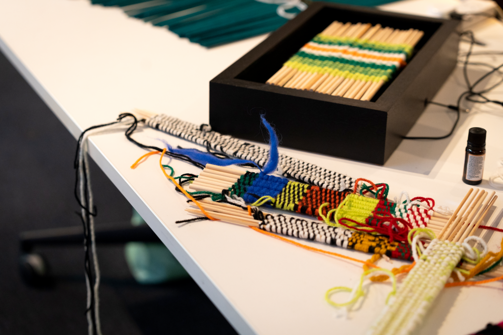 Small woven tapestries on top of a table next to a black box holding another tapestry. The tapestries are made from brightly colour threads and are woven around small wooden sticks.