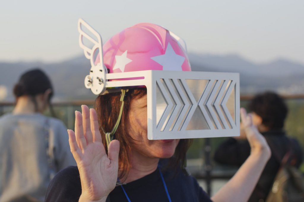 A person holding their hands up, whilst hey are wearing a pink helmet. The helmet has white stars and a silver and white visor that is in front of their face. 
