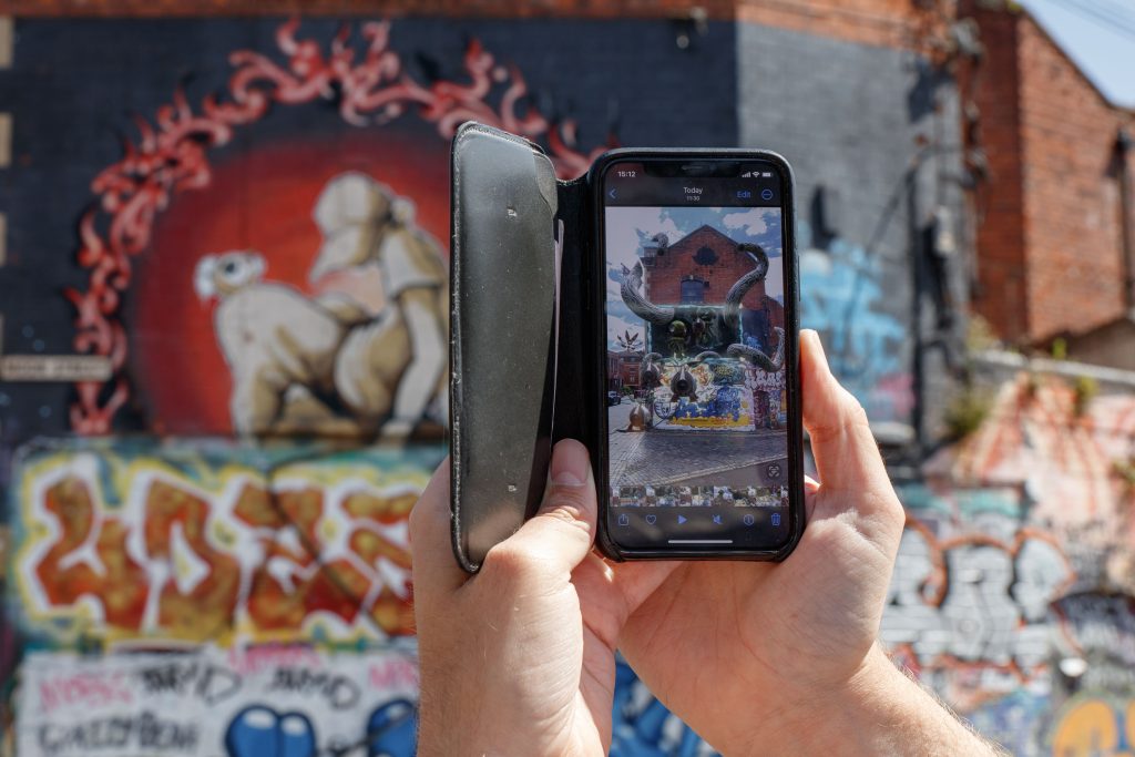 Hands holding up a mobile phone in front of a mural. On the phone an augmented reality experience shows aliens emerging from the mural. 